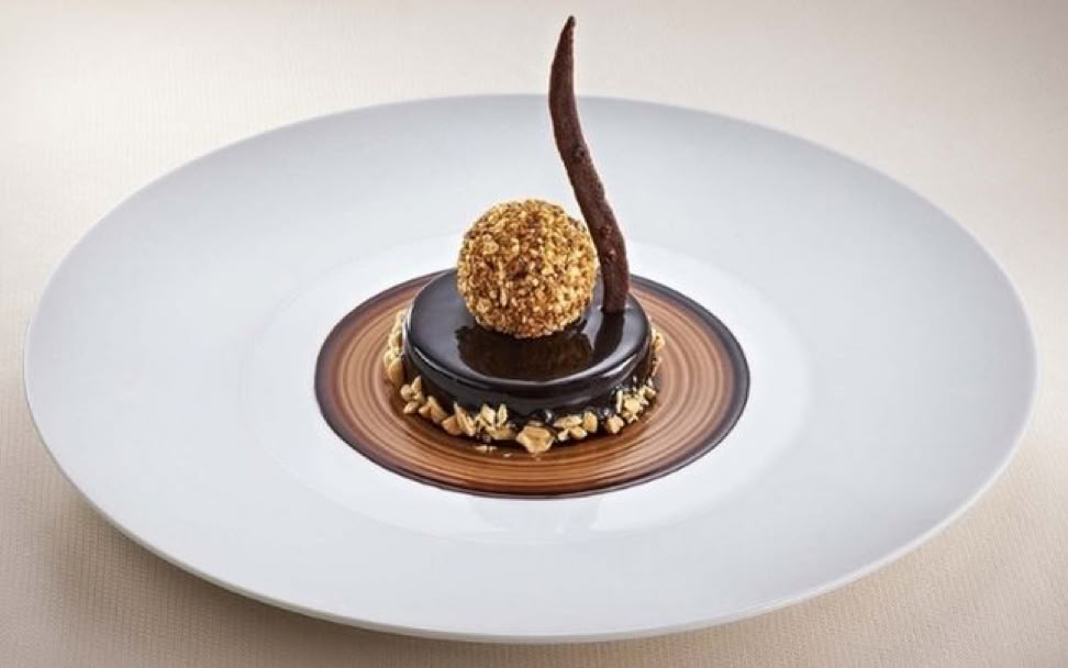 Ein Dessert auf einem weißen Teller, bestehend aus einer runden Schokoladenbasis mit einer goldenen Kugel und einer dekorativen Schokoladenspirale.
