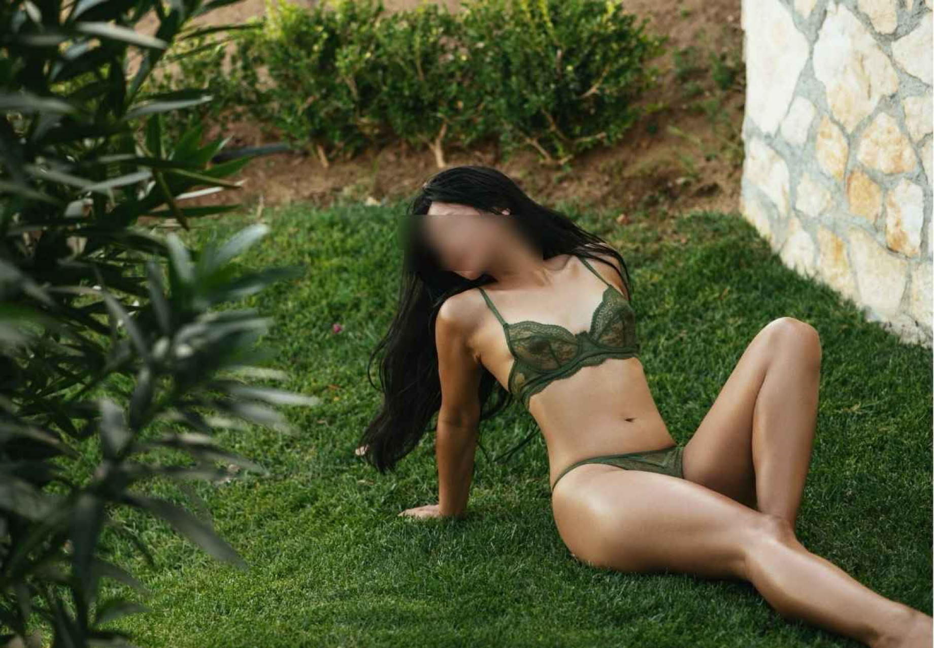 Eine Person in grüner Unterwäsche sitzt auf Gras neben einer Steinmauer.